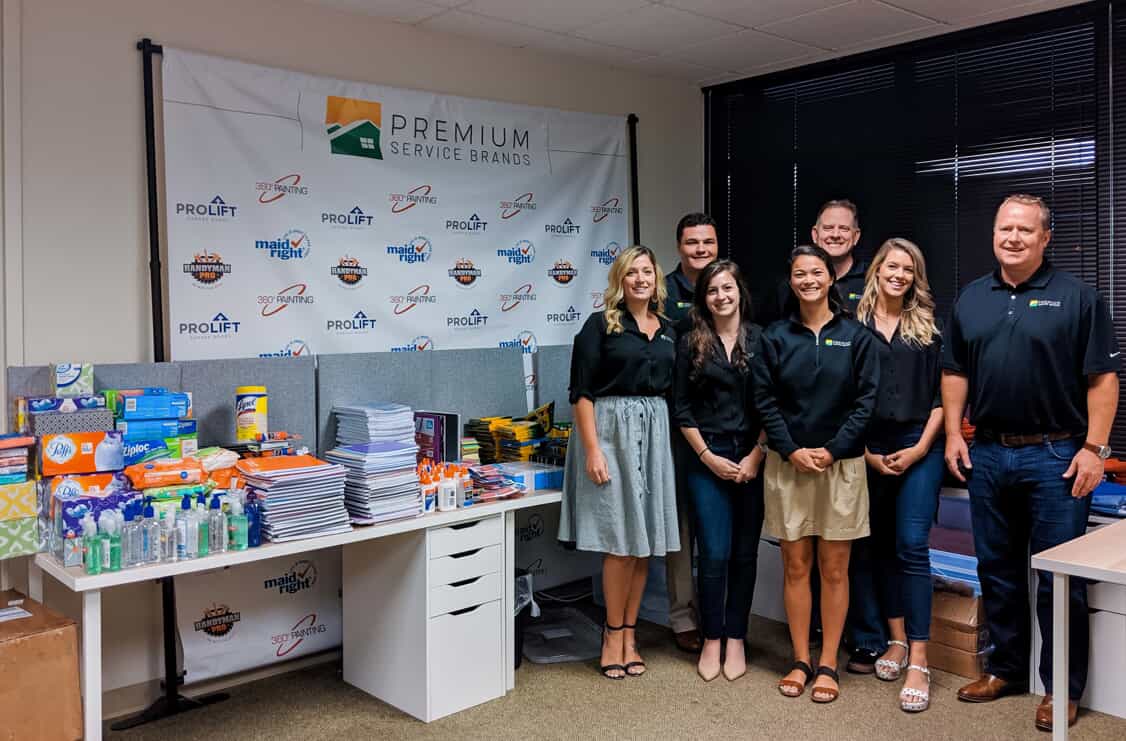 Several people stand before school supplies arranged on a desk for a charity drive